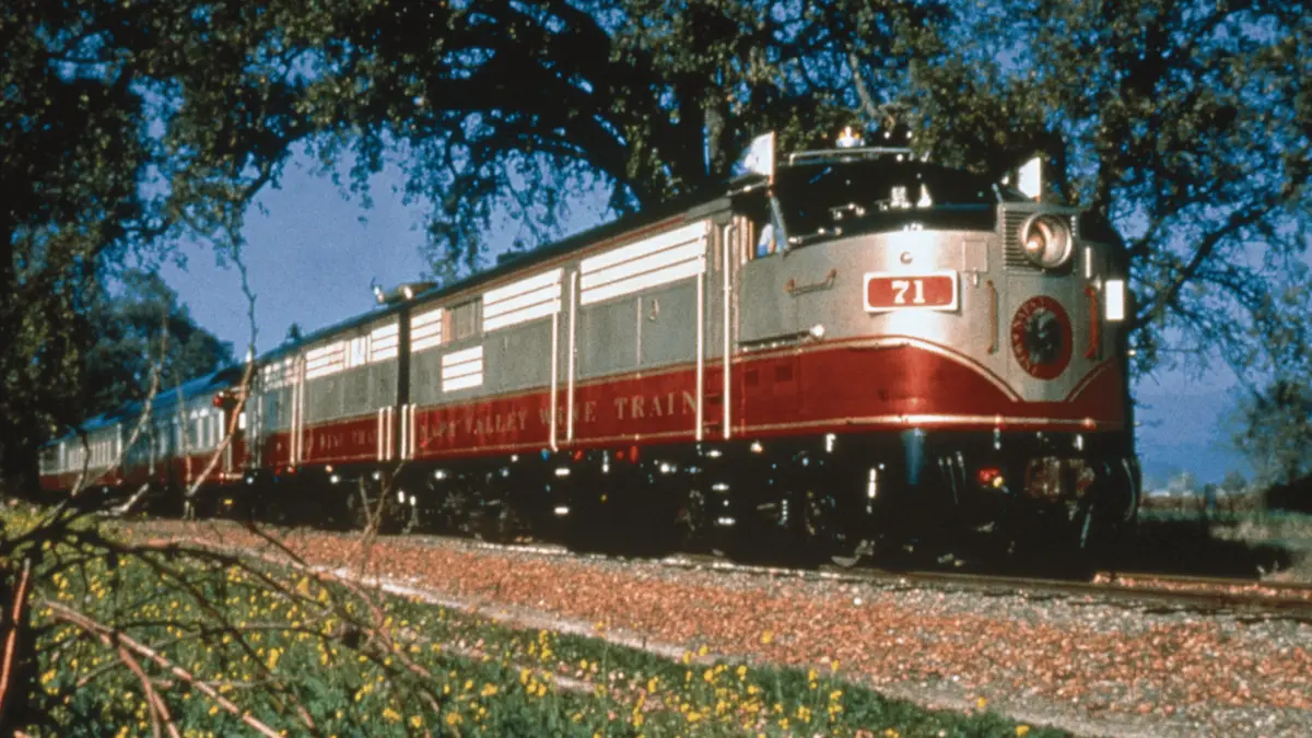 A vintage red and silver passenger train with the number 71 on the front travels on a track through a rural area, surrounded by trees and foliage. The clear sky and sunlight highlight the train's streamlined design, reminiscent of a picturesque Napa Valley visit.