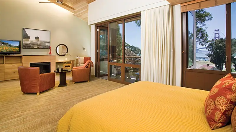 A spacious room in one of the best group hotels in North Bay features a cozy bed with a yellow bedspread, wooden furniture, a flat-screen television, armchairs around a small table, a fireplace, and large windows with a view of the Golden Gate Bridge in the distance.