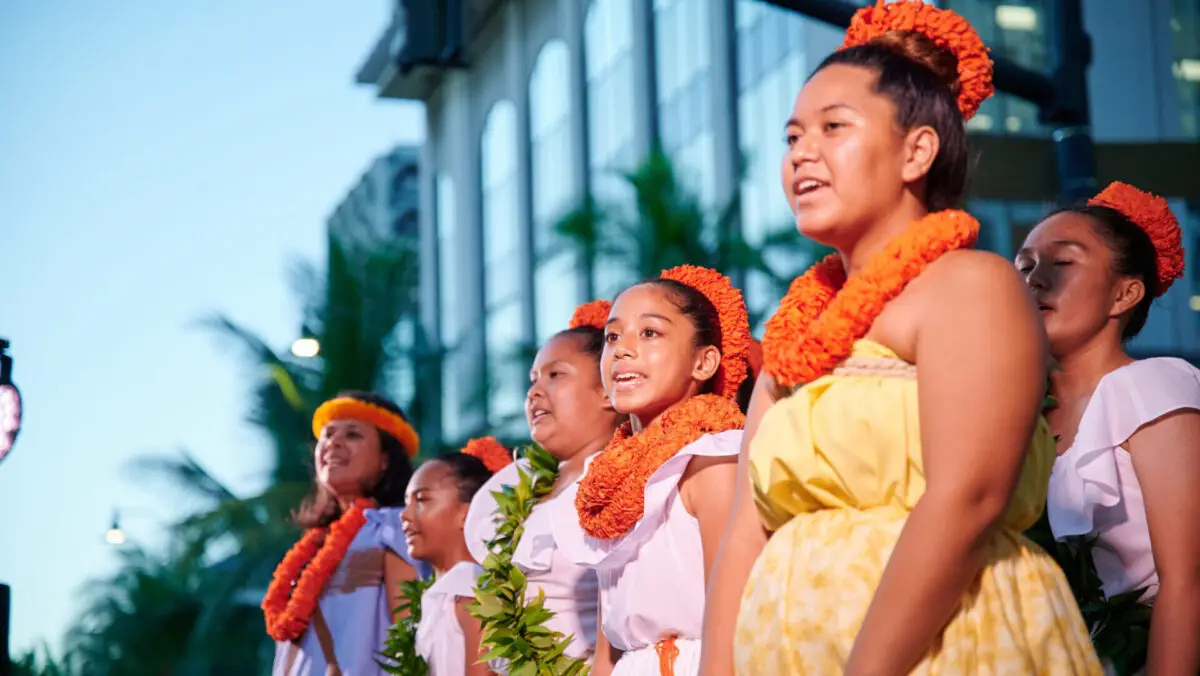 A group of people dressed in traditional Hawaiian attire sings together outdoors at an annual event in Waikiki. They wear vibrant orange leis and headbands, positioned in front of a building with tall windows and palm trees, under a clear sky.