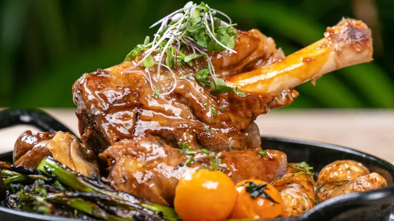 A close-up of a succulent, glazed lamb shank garnished with fresh herbs. The dish is served in a black skillet, accompanied by grilled vegetables and cherry tomatoes, presented against a blurred green background reminiscent of Annual Events Waikiki.