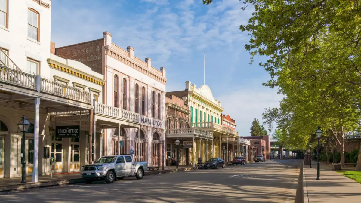 A quiet, sunny street in a historic downtown area offers a glimpse of what to do in Northern California. The street is lined with old-fashioned buildings featuring vintage architecture and balconies. Cars are parked along the side, and leafy green trees line the right side of the street. The sky is clear.