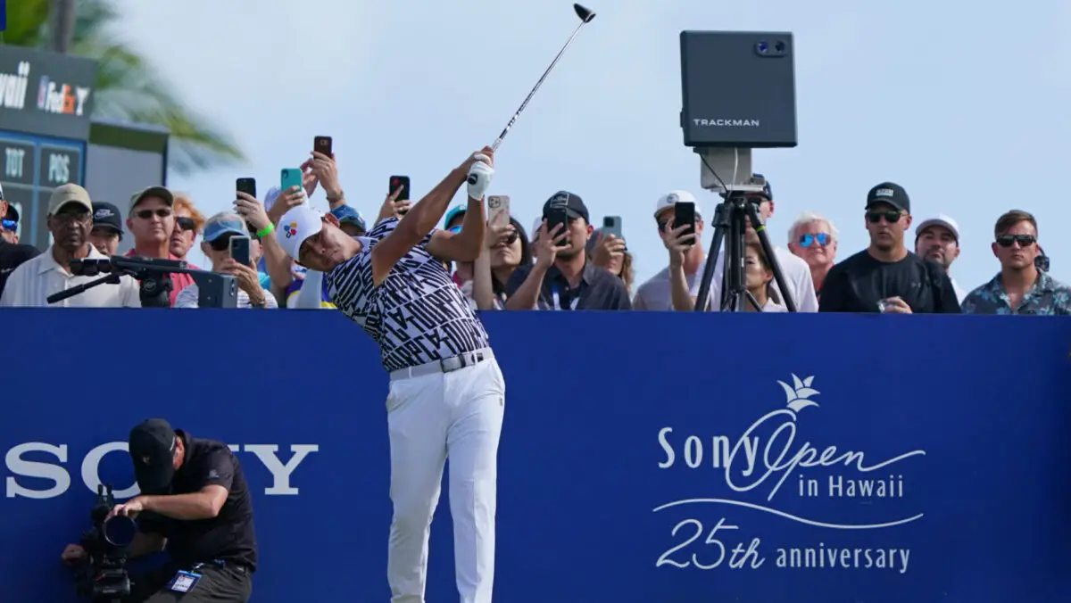 A golfer in a patterned shirt and white pants takes a swing at the Sony Open in Hawaii, an annual event celebrating its 25th Anniversary. Spectators capture the moment on their phones beneath a sign that reads "Sony Open in Hawaii," set against the stunning backdrop of Waikiki.