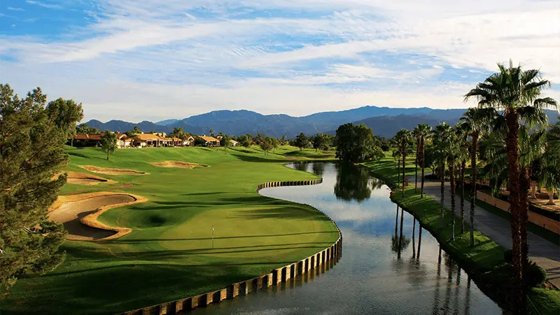 A scenic golf course under a blue sky with scattered clouds. The course features well-maintained greens, sand bunkers, and a winding water hazard lined with palm trees. In the background, there are houses and rolling hills, with distant mountains beyond—truly one of the best places to visit in Palm Springs.