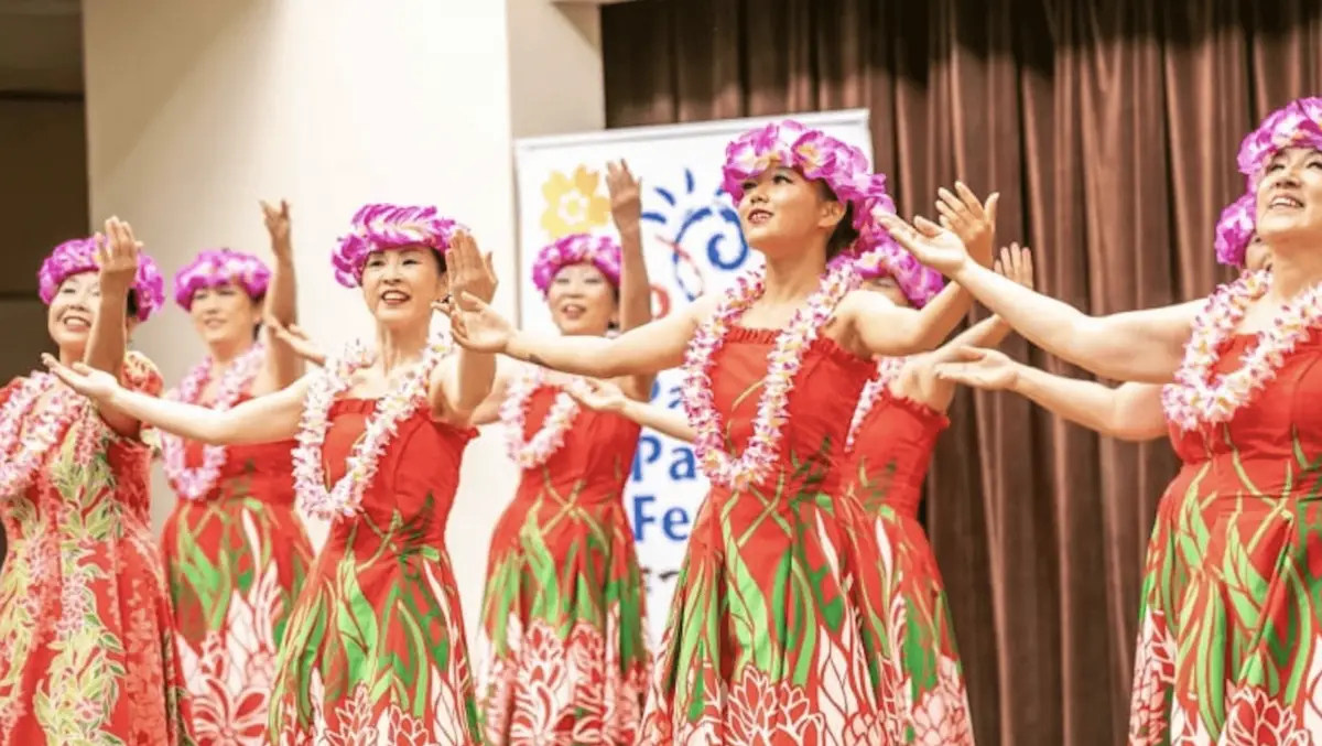 A group of women dressed in red dresses with green leaf patterns and purple flower crowns perform a hula dance, celebrating Hawaiian holidays. They are in mid-motion, smiling and extending their arms. In the background, there is a brown curtain and a partially visible sign.