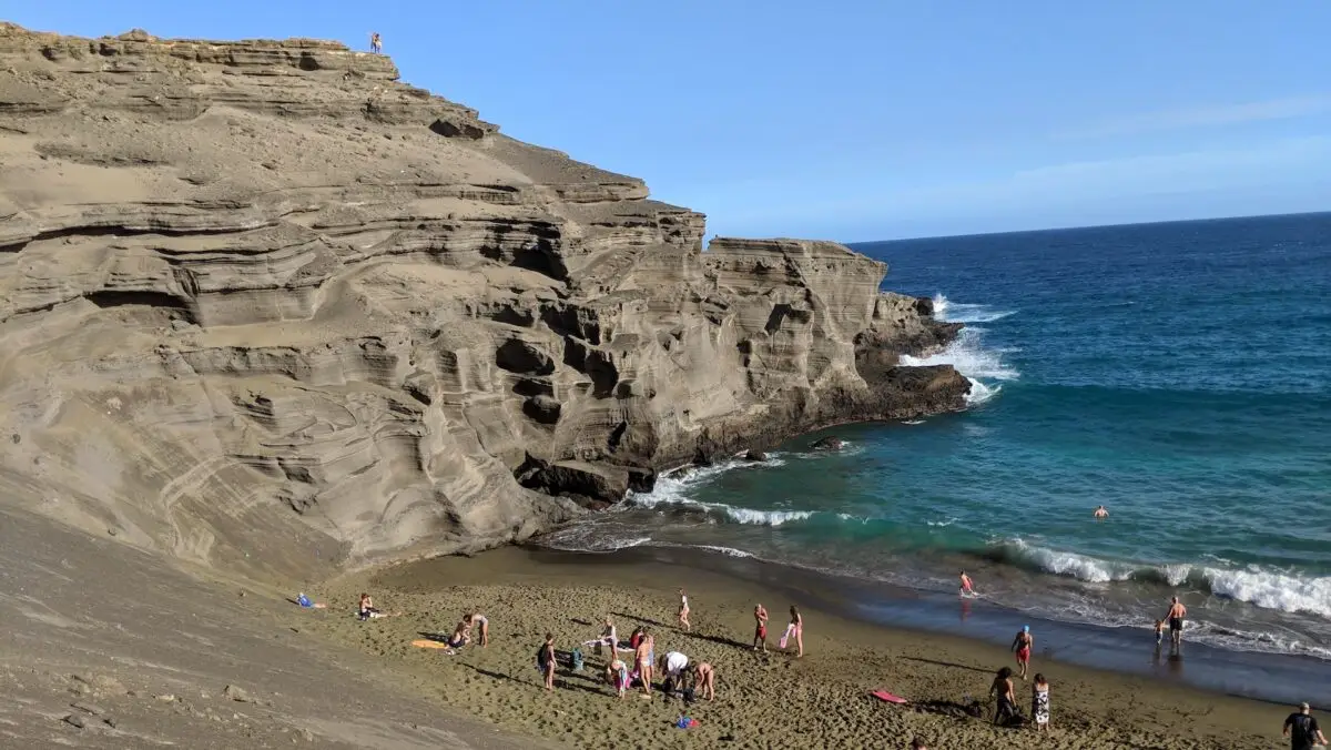 A picturesque beach scene with a large rocky cliff on the left, people sunbathing and playing on the sandy shore of Papakolea Green Sand Beach, and others swimming in the clear blue ocean. The sky is clear and bright, creating a serene and inviting environment.