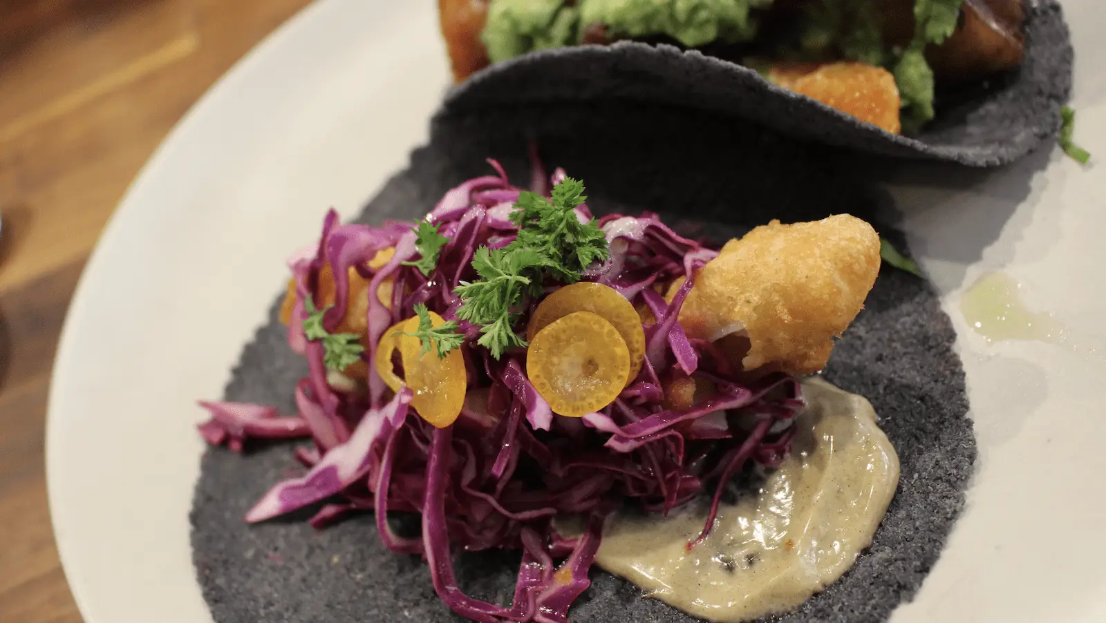 Close-up of a taco with a black tortilla, filled with deep-fried fish, shredded purple cabbage, small orange slices, and a dollop of light-colored sauce. Fresh herbs are placed on top as garnish. Another similar taco is partially visible in the background.