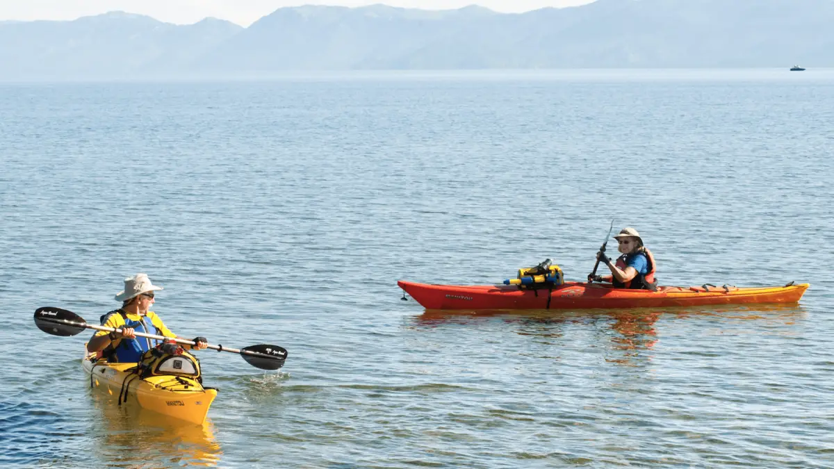 Two people in kayaks enjoy a calm, sunny day on Lake Tahoe, with a mountain range visible in the background. One person paddles a yellow kayak, while the other holds a fishing rod in an orange kayak. Both are wearing hats and safety vests—prime examples of outdoor activities Tahoe has to offer.