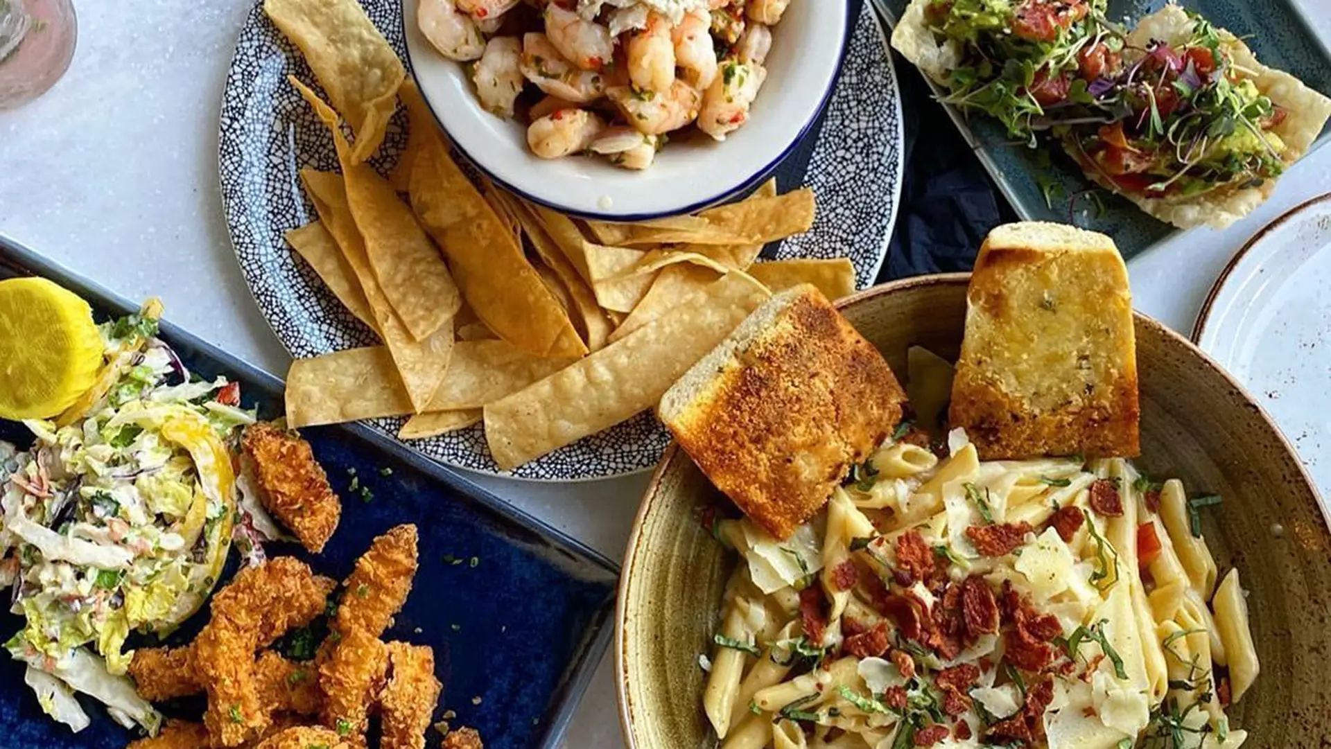 A spread of the best lunch Santa Barbara has to offer, featuring pasta topped with bacon bits and garlic bread, fried chicken strips, shredded coleslaw, a shrimp dish with tortilla strips, and an avocado boat filled with fresh greens and diced tomatoes.