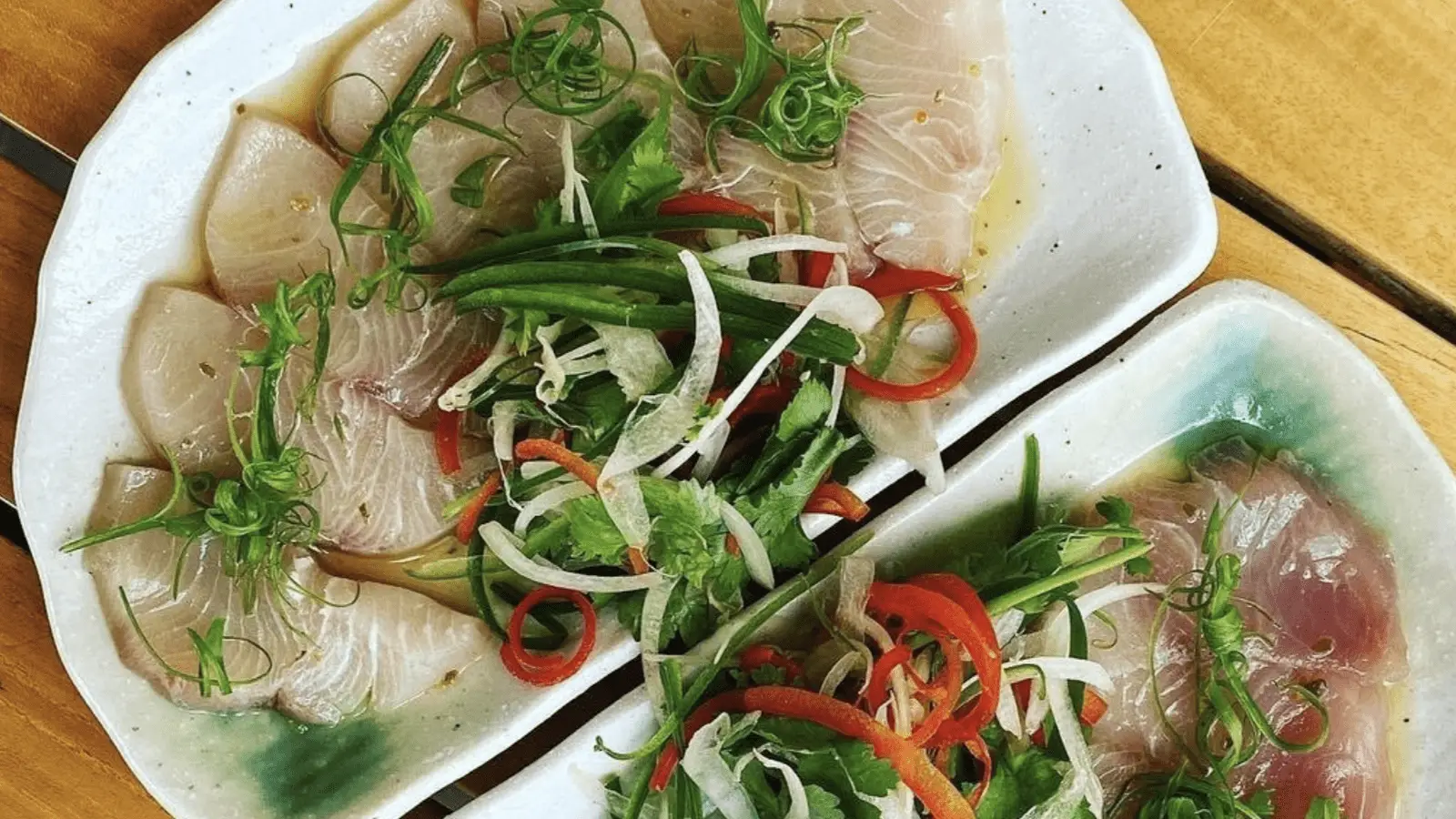 Close-up of three white rectangular plates containing thin, translucent slices of sashimi, garnished with sliced green onions, red chili peppers, and other fresh greens. The plates are placed on a wooden surface, presenting the best dinner Ko Olina has to offer.