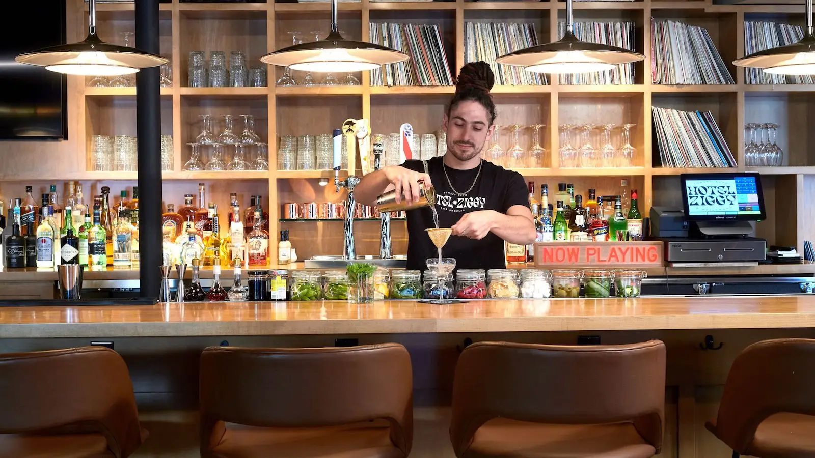A bartender with a high bun hairstyle mixes a drink at Hotel Ziggy's modern bar, featuring a wooden counter and shelves stocked with bottles, glasses, and vinyl records. The counter has various herbs, fruits, and jars. A neon sign reads "NOW PLAYING" in red. Four barstools line the counter.