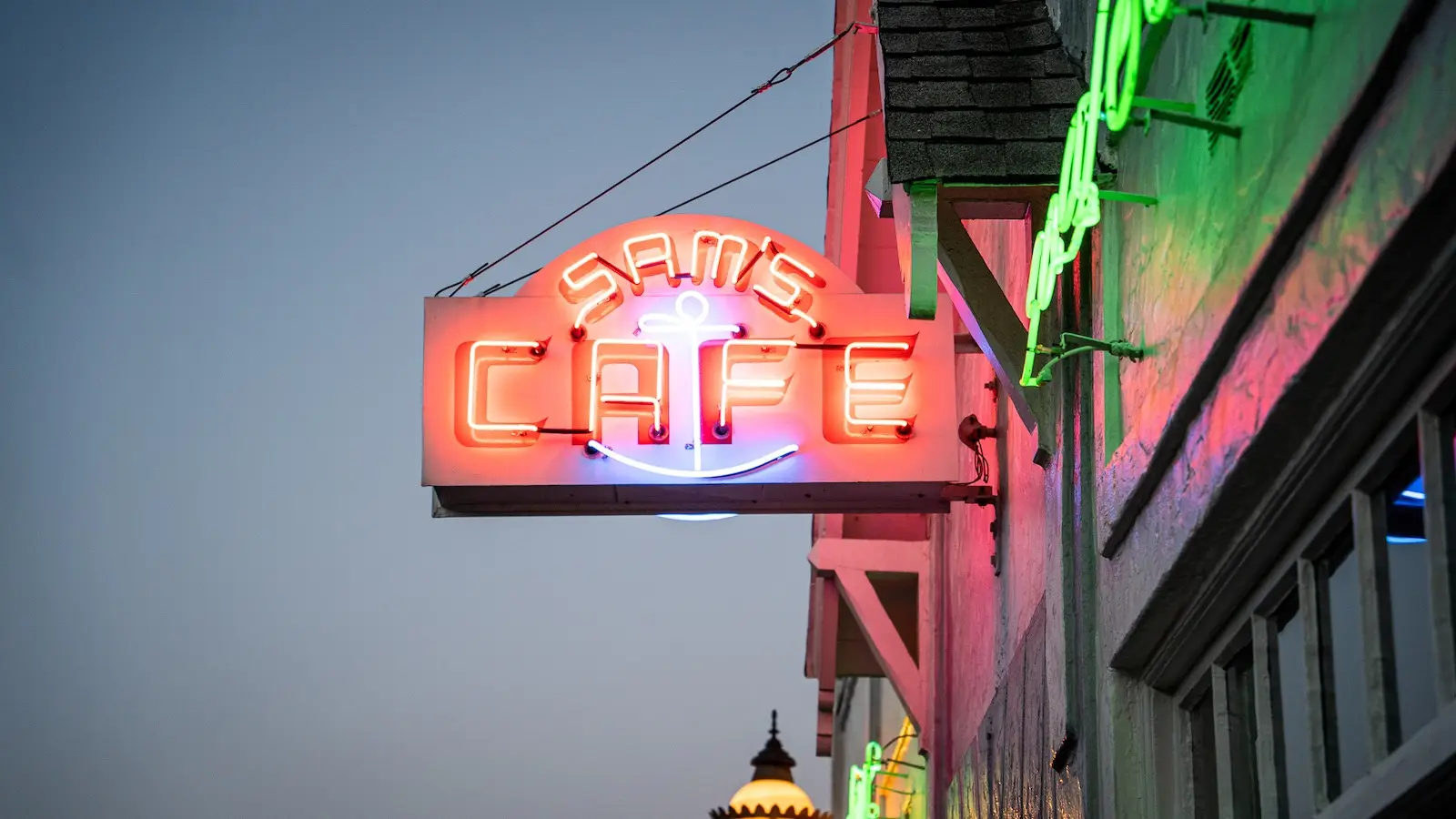 A glowing neon sign that reads "SAM'S CAFE" with an anchor symbol below is mounted on the exterior of a building in Tiburon. The sign is illuminated in pink and red, contrasting with the dusky evening sky. Other neon signs and part of a building's roofline are visible in the background.