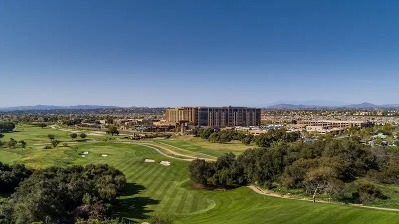 Aerial view of a large multi-story building complex surrounded by an expansive, lush green golf course. The landscape includes trees, sand traps, and well-manicured fairways where you can enjoy a day of golfing, all set against distant mountains and a clear, blue sky in the background.