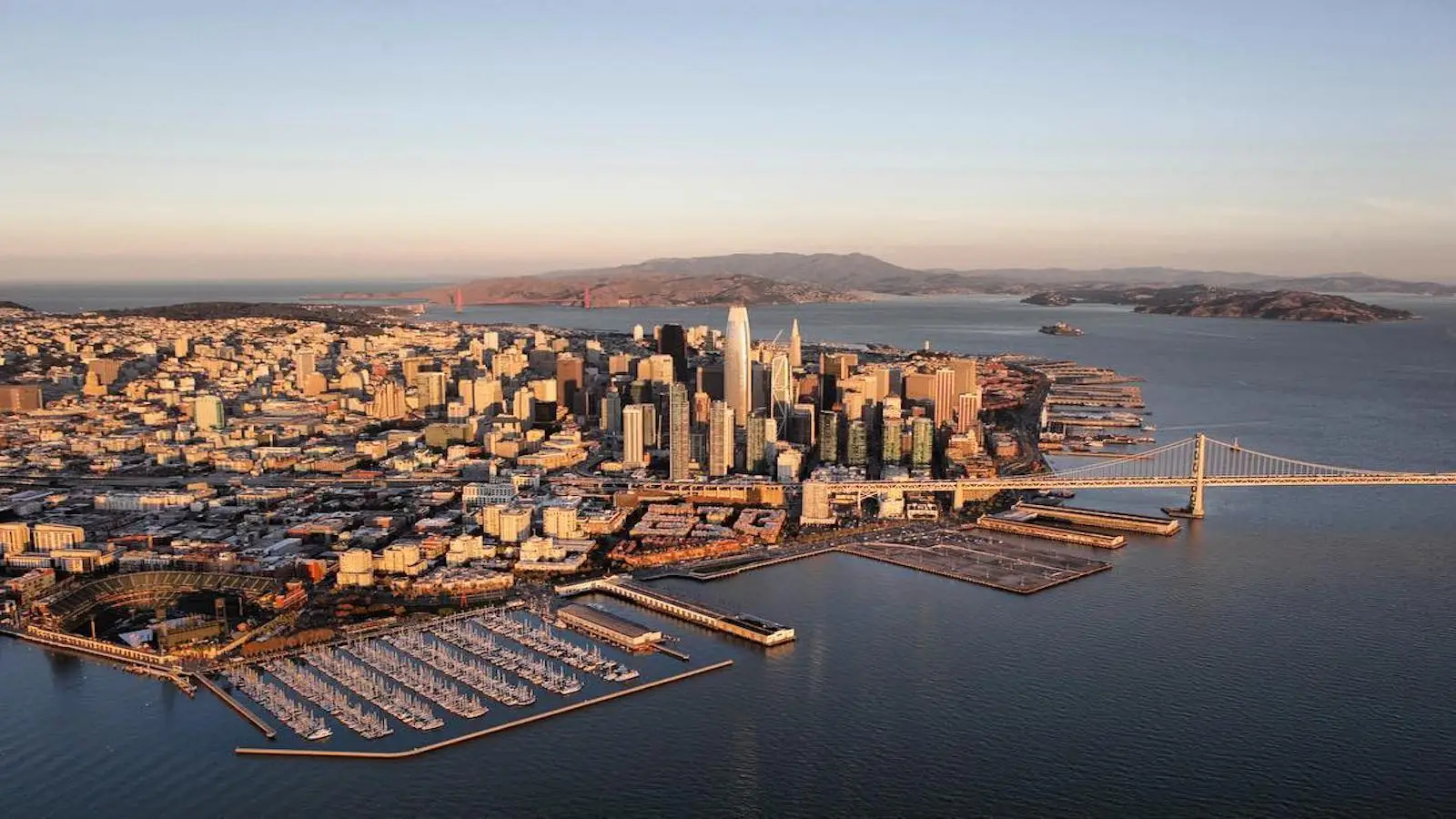 Aerial view of San Francisco's downtown skyline with the Golden Gate Bridge in the background. The image shows the Bay Bridge to the right and a marina with numerous boats docked in the foreground, perfect for local getaways, with the city extending outwards on a sunny day.