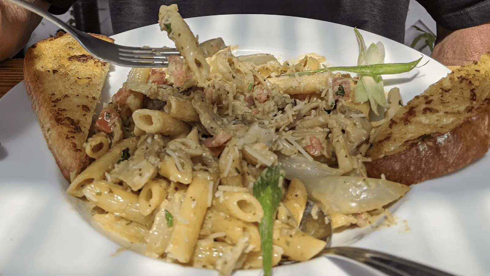 A close-up of a person holding a fork digging into a plate of creamy penne pasta garnished with herbs and bits of what appears to be bacon or ham, arguably the best lunch on the Kohala Coast. Two crispy garlic bread slices are placed on the rim of the white plate.