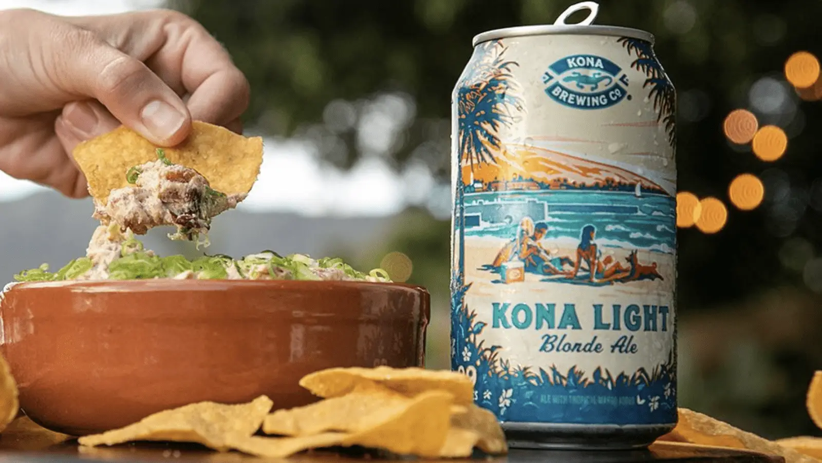 A hand dips a chip into a bowl of dip beside a can of Kona Light Blonde Ale, perfect for the best lunch. The can's design features two people lounging on a beach with palm trees and an ocean backdrop. Loose chips and a blurred, outdoorsy background complete the scene.