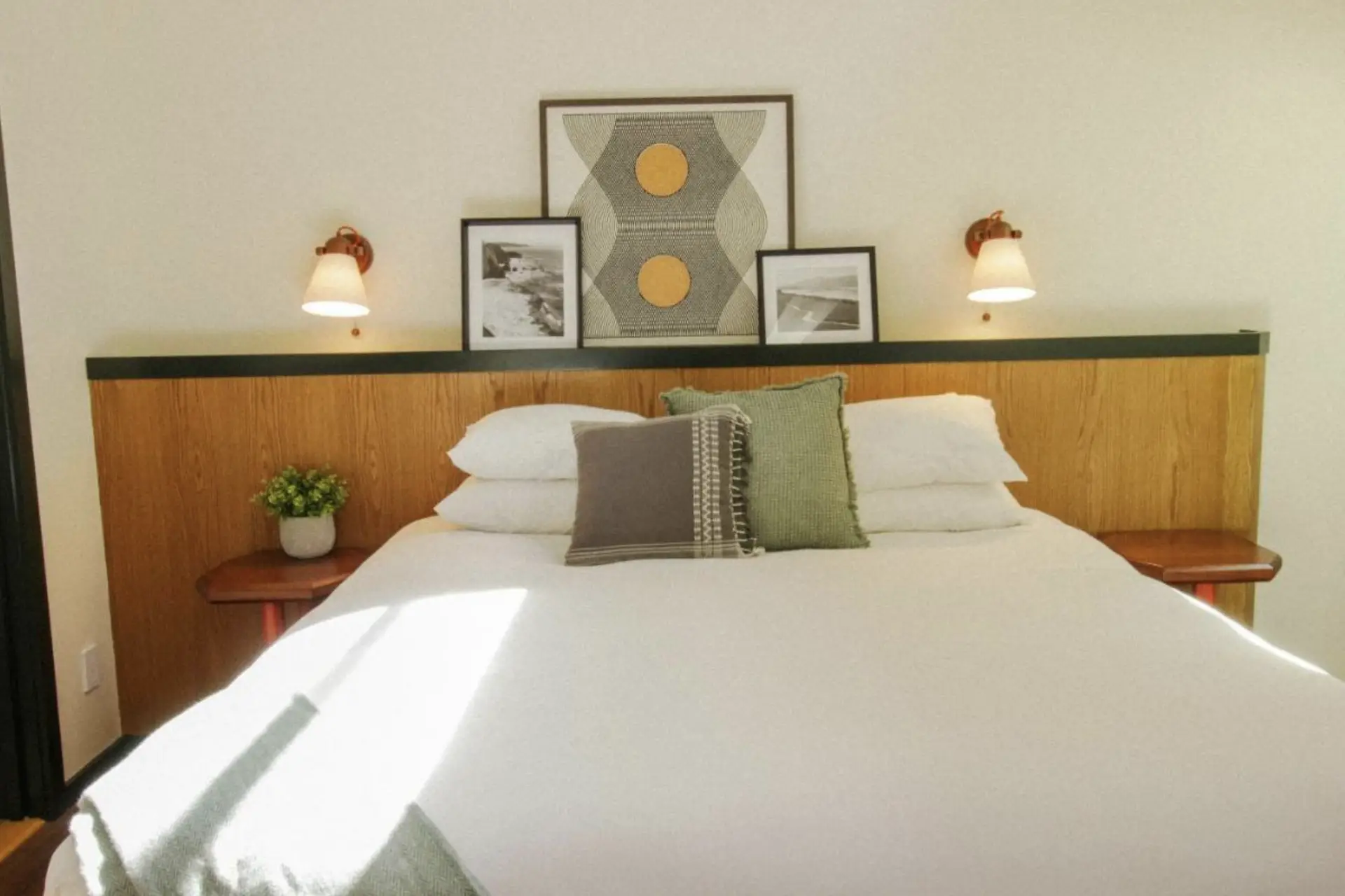 A neatly made bed with white linens, green and gray accent pillows, and a minimalistic wooden headboard. The headboard features built-in nightstands, small potted plant decor, framed art, and two sconces with warm lighting. Sunlight streams in, brightening the room—a scene reminiscent of the best group hotels North Bay has to offer.