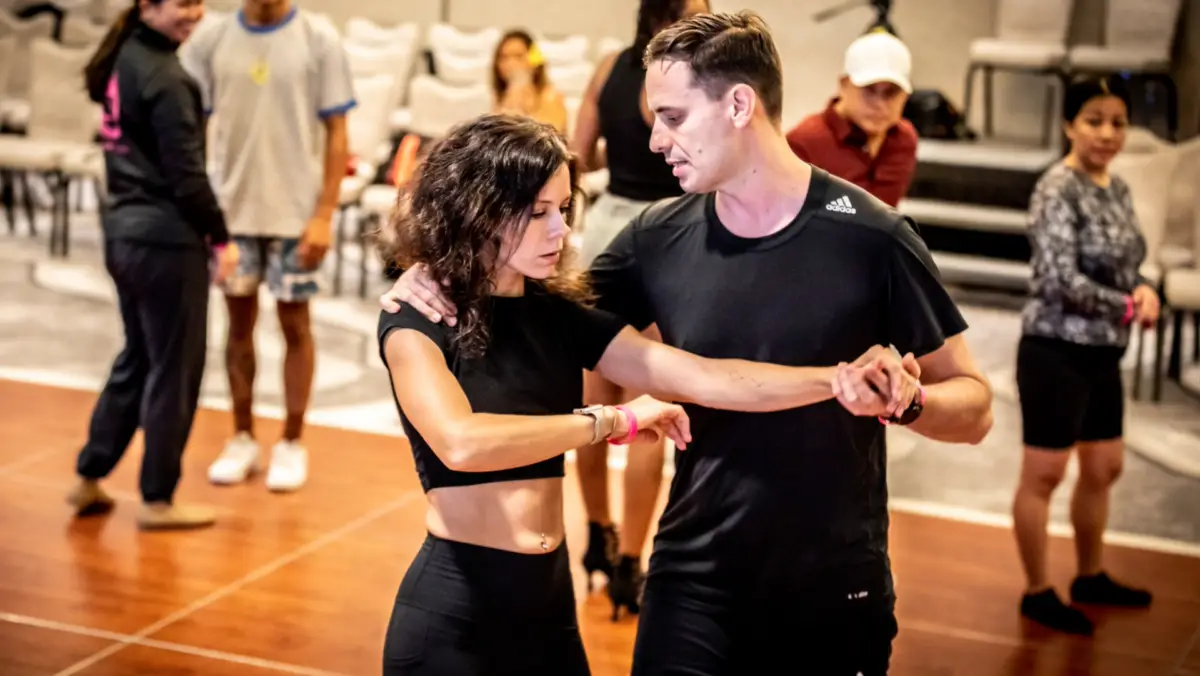Two people, a man and a woman, dance closely together in a well-lit indoor space. Both wear black outfits and are focused on their movements. The woman has short curly hair and a cropped top, while the man holds her hand and shoulder. Other individuals are visible in the background, reminiscent of annual events in Waikiki.