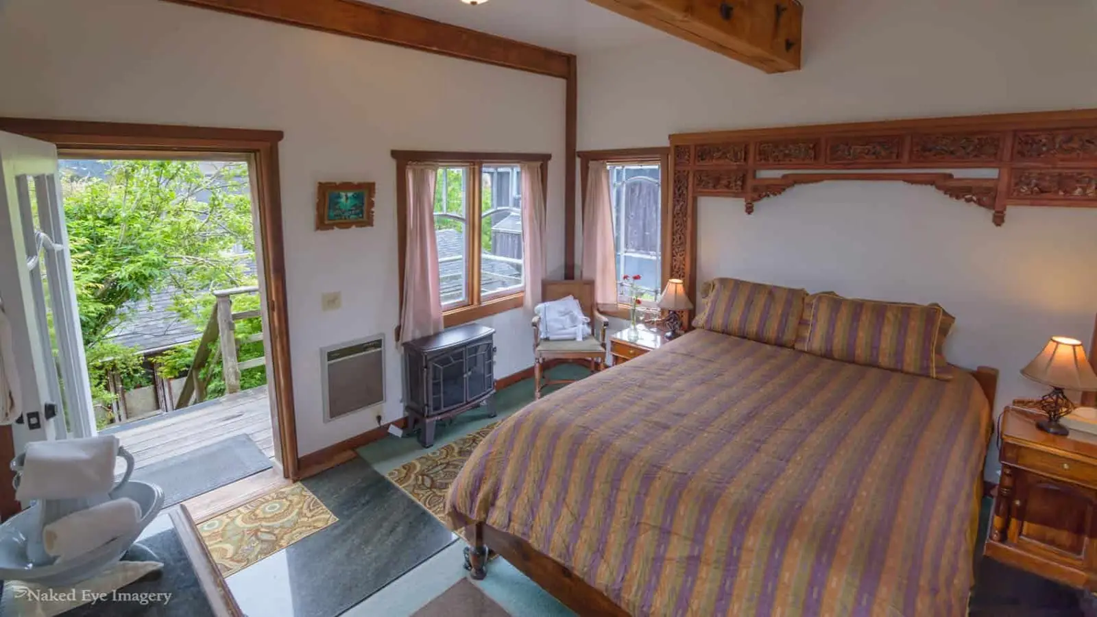 A cozy bedroom in one of the best wellness hotels North Bay features a large wooden bed with a striped comforter, flanked by traditional carved wooden headboards. The room has panoramic windows, a door leading to an outdoor deck, and various furnishings including a nightstand, chairs, and a small heating unit.