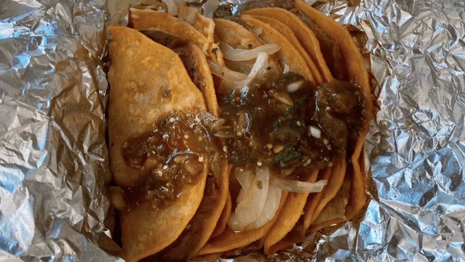 A batch of folded tacos filled with meat, topped with onions and green salsa, placed on a sheet of aluminum foil. The tacos are warmly colored and appear to be freshly cooked, each taco exuding a mouth-watering aroma.