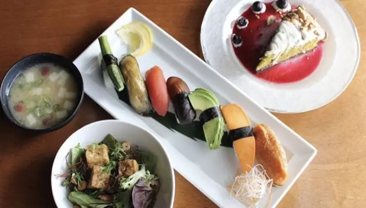 An assortment of vegan sushi pieces arranged on a white rectangular plate, accompanied by a bowl of salad with tofu, a small bowl of miso soup, and a dessert plate featuring a slice of cake with blueberries and sauce. This delightful spread would fit right in at one of the top vegan restaurants in Oahu. The meal is set on a wooden table.