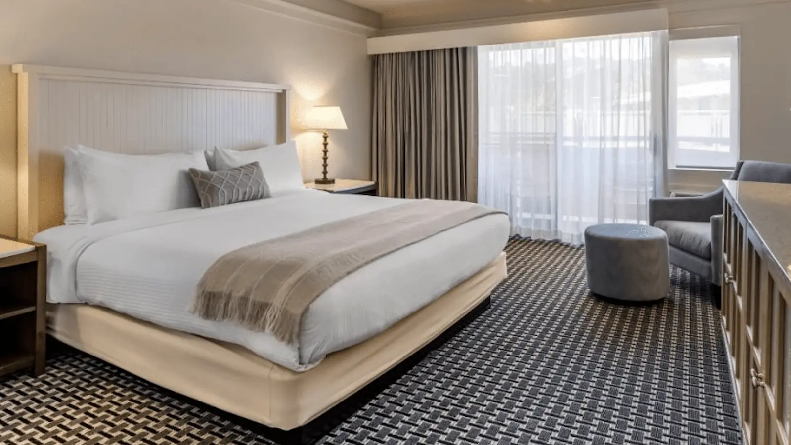 A Marin County hotel room with a large bed adorned with white linens and a beige throw blanket. To the right, there's a window with sheer curtains, a grey armchair, and an ottoman. A bedside lamp is on the nightstand to the left, and the room features patterned carpeting.