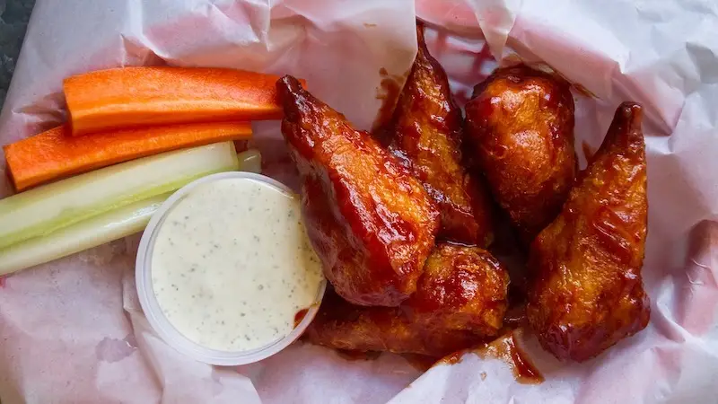 A serving of glazed chicken wings is placed on parchment paper, accompanied by carrot sticks, celery sticks, and a small cup of ranch dressing. The wings appear to be coated in a dark, sticky sauce—a stark contrast to the vibrant veggie options seen in top vegan restaurants Oahu offers.