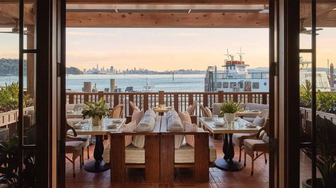 The open-air restaurant deck boasts wooden tables and cushioned seating, all facing a scenic waterfront view reminiscent of Tiburon. Nearby boats are docked with a city skyline visible in the distance under a light sunset sky, while indoor plants frame the foreground.