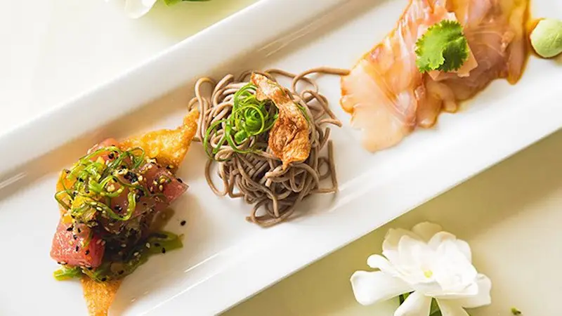 A rectangular white plate presents the best lunch on the Big Island with three gourmet dishes arranged in a row. On the left, seared tuna with seaweed; in the center, soba noodles topped with sliced scallions and a crispy garnish; on the right, thinly sliced sashimi garnished with cilantro. White flowers decorate the table.