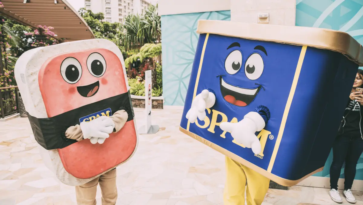 Two cheerful mascots in costume, one as a can of SPAM with a blue and yellow label and the other as a piece of pink SPAM meat with a black label, stand outside amidst greenery and buildings. Celebrating Hawaiian holidays, they bring smiles while someone snaps photos nearby.
