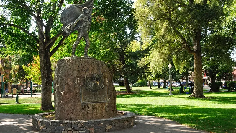 A park with green grass and large trees, featuring a prominent statue of a man holding a large flag on a stone pedestal. The sunny area includes park benches, pathways, and a few people in the distance—an ideal spot to explore wine country and enjoy outdoor activities.