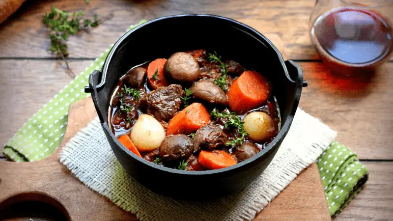 A cast iron pot filled with a hearty beef stew, featuring chunks of beef, carrots, onions, and mushrooms. The pot is resting on a burlap cloth and wooden cutting board, with a glass of red wine and a sprig of thyme in the background—a perfect addition to your winter menu.