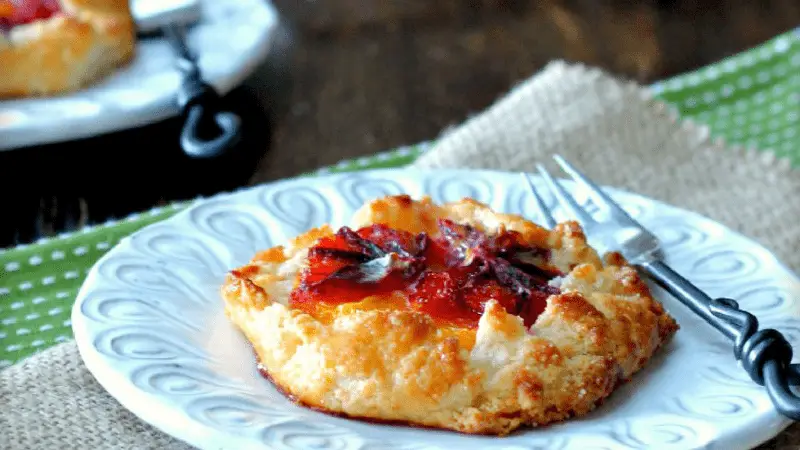 A close-up of a fruit tart on a white decorative plate with a fork beside it. The tart has a flaky, golden crust and a red fruit filling, possibly strawberry or raspberry—perfect for the winter menu. Another plate with a similar tart is partially visible in the background.