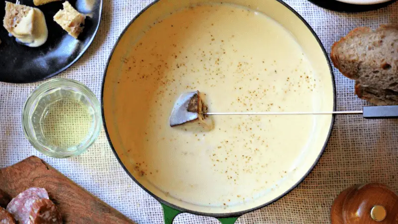A pot of cheese fondue with a piece of bread dipped on a fondue fork. Surrounding the pot are slices of bread, a wooden board with salami, a glass of white wine, and a plate with additional pieces of bread. Perfect for any winter menu, the scene is set on a white tablecloth.