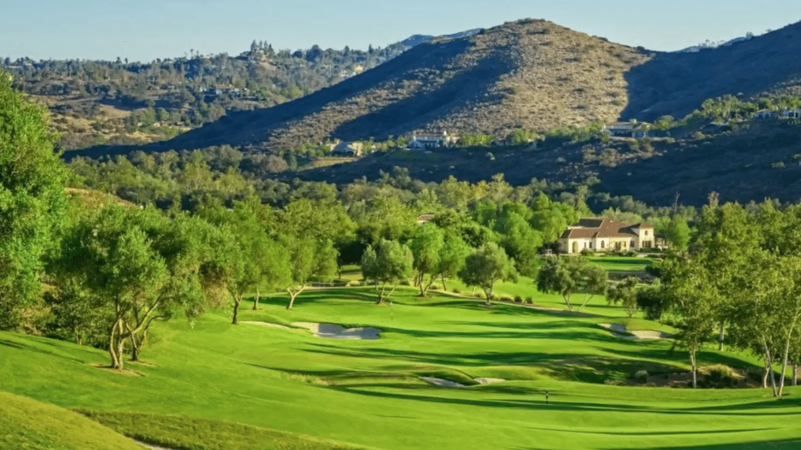 A well-maintained golf course with bright green fairways and sand bunkers sits nestled among rolling hills with scattered trees and houses. The picturesque backdrop includes lush vegetation and distant mountains under a clear blue sky, making it one of the best golfing experiences in San Diego.