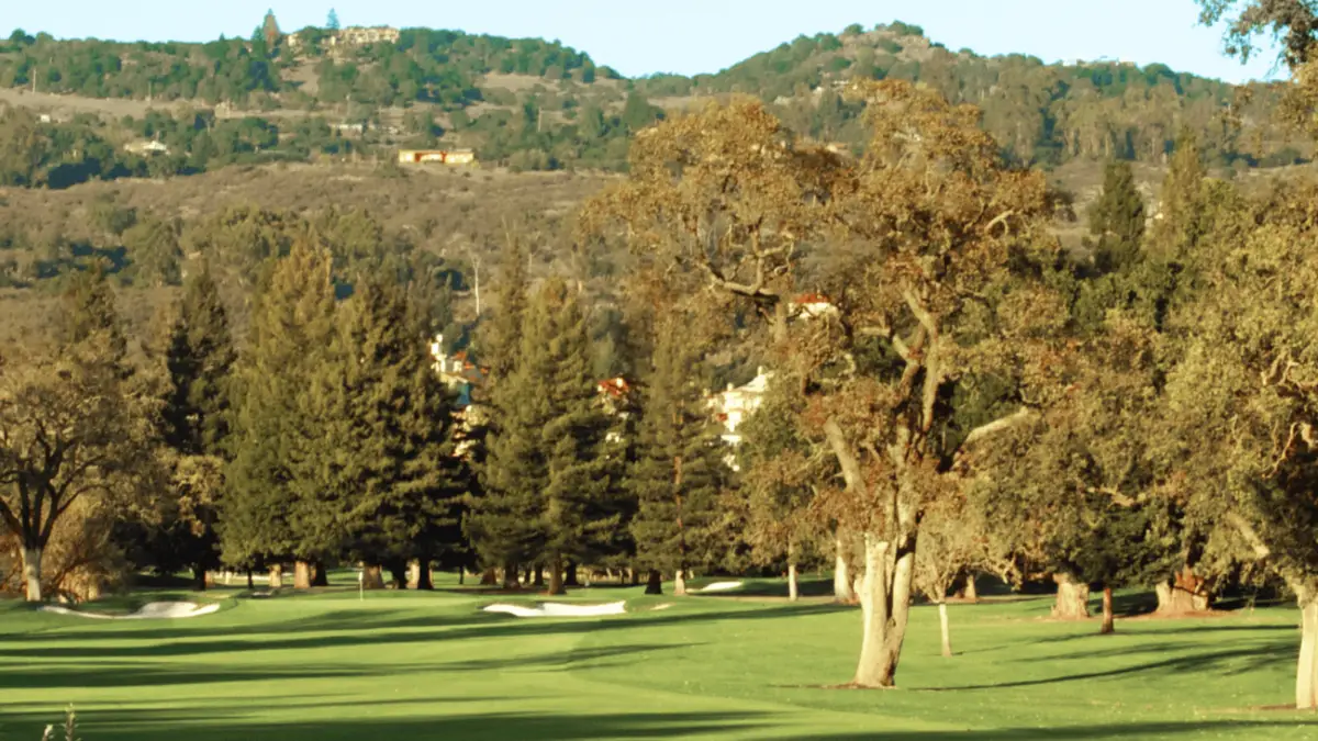 A scenic view of an intercontinental golf course with lush green grass, sand traps, and tall trees. Hills covered in trees and some houses are visible in the background under a clear blue sky. The foreground features a few large, shadowed trees.