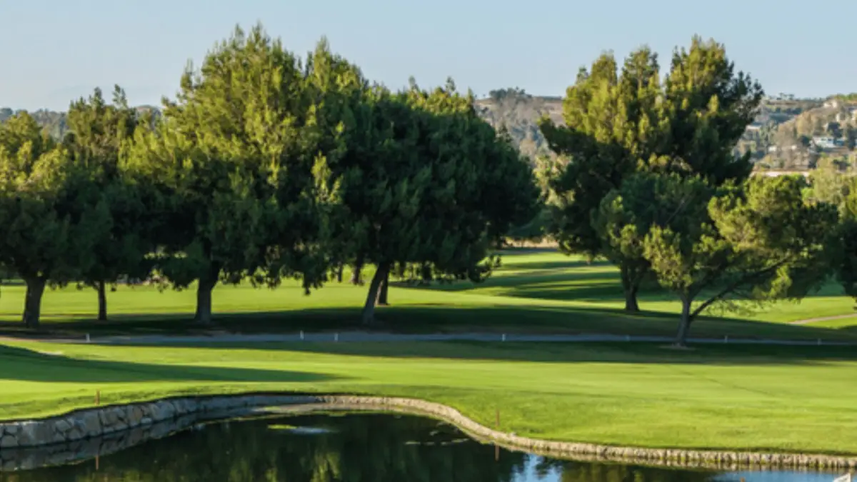 A scenic view of a golf course featuring lush green grass, large trees providing shade, and a calm pond in the foreground. The sky is clear and blue, with distant hills visible in the background—truly one of the best golf courses Bay Area enthusiasts will appreciate.