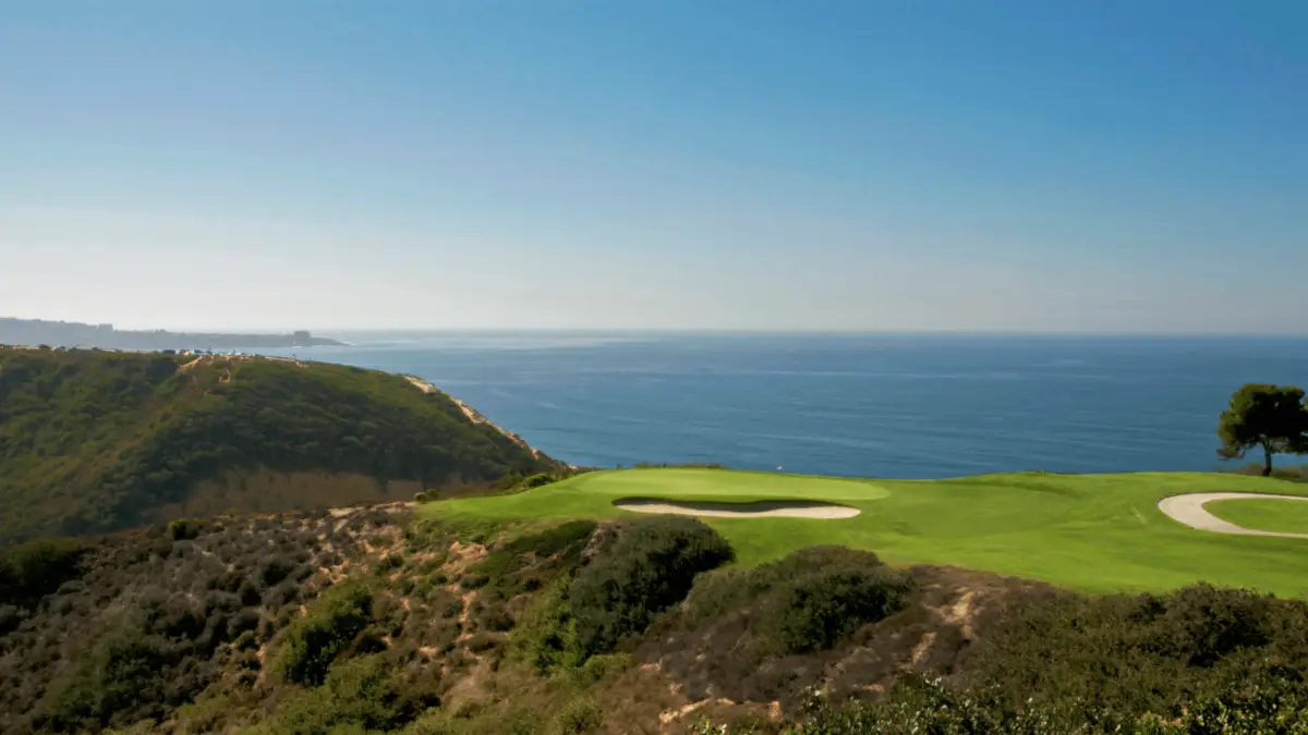 A picturesque golfing course overlooking the ocean features a pristine green with sand bunkers on a cliffside. The lush green contrasts with the rugged terrain and the deep blue sea under a clear sky, making it one of the best golf courses in the Bay Area for things to do in San Diego.