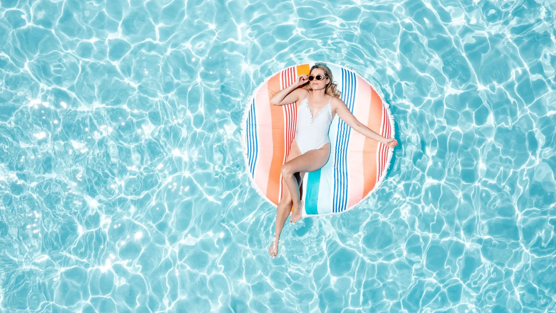 A person relaxes on a colorful striped inflatable float in a swimming pool during their wine country getaway. The individual is wearing sunglasses and a white swimsuit, lying back with one arm resting on the float and the other hand touching the clear, sparkling water as sunlight dances across the surface.
