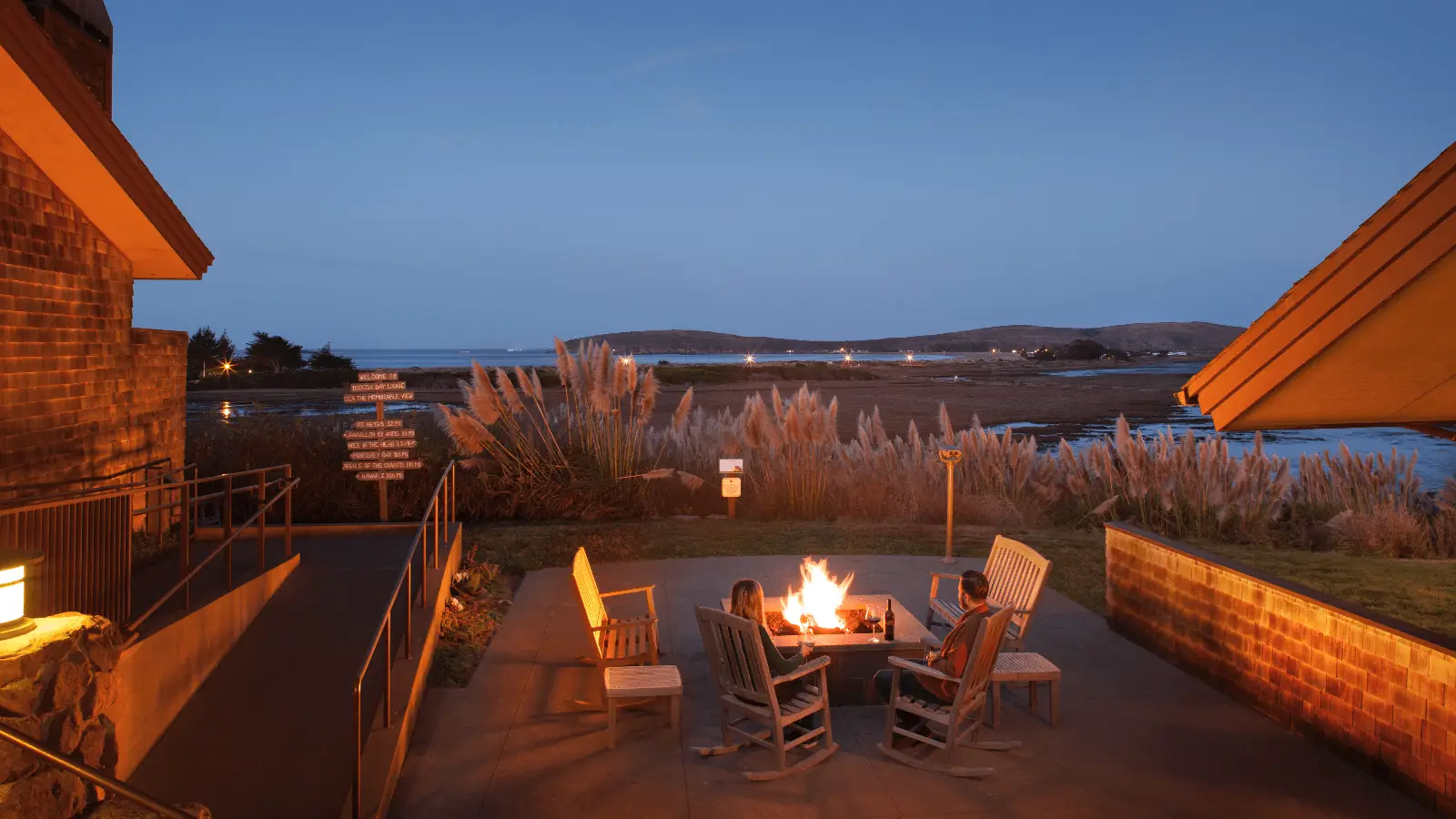 A group of chairs surrounds a fire pit on a patio overlooking a serene body of water at dusk. This perfect local getaway is framed by two buildings, with lush grasses and distant hills visible in the background. The sky is clear, transitioning from light to dark blue.