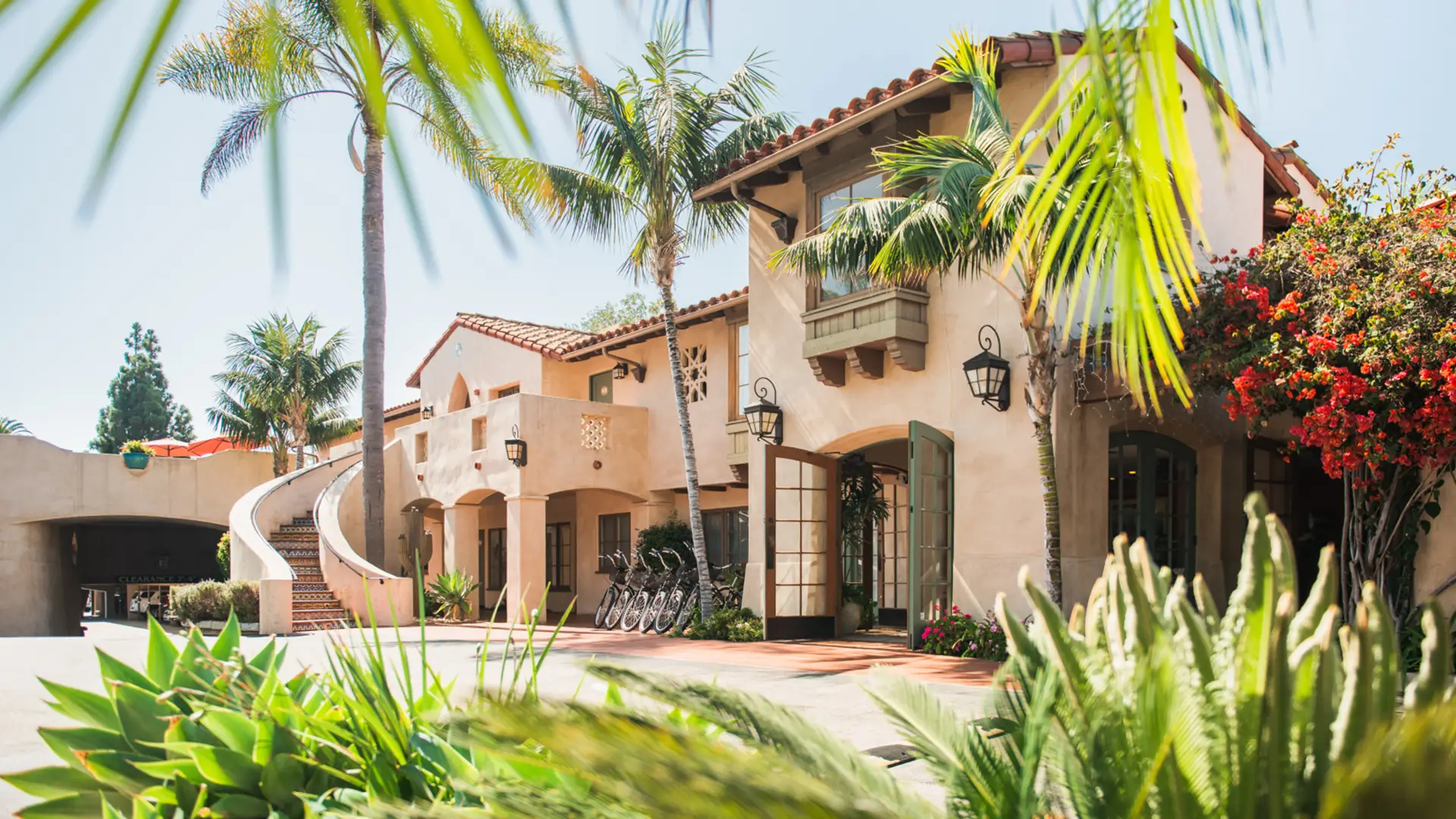 A large, Spanish-style villa with a terracotta tiled roof, arched doorways, and a spiral staircase is surrounded by lush tropical plants and palm trees. Bicycles are parked near the entrance, and vibrant bougainvillea adds a pop of color. The sky is clear and blue—truly one of the best group getaway hotels Santa Barbara has to offer.