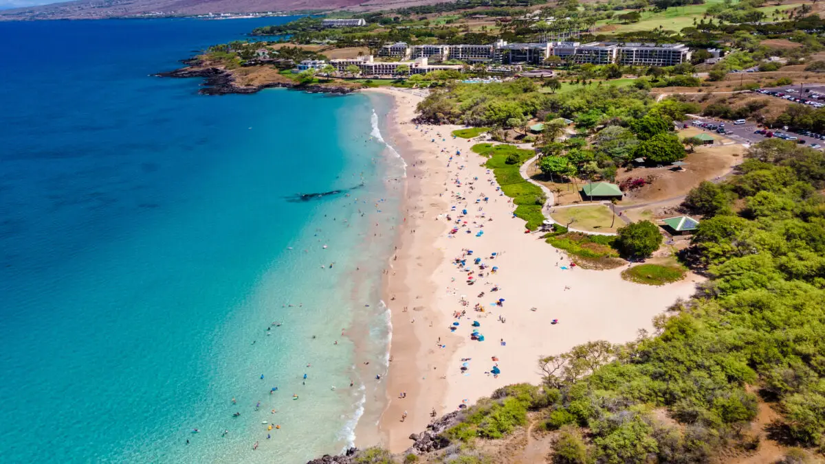 Aerial view of a sunny Hapuna Beach with turquoise waters, lined with sunbathers and umbrellas. Behind the beach are lush green areas, a few structures, and a parking lot. The coastline extends to the horizon, with buildings and hills in the background.