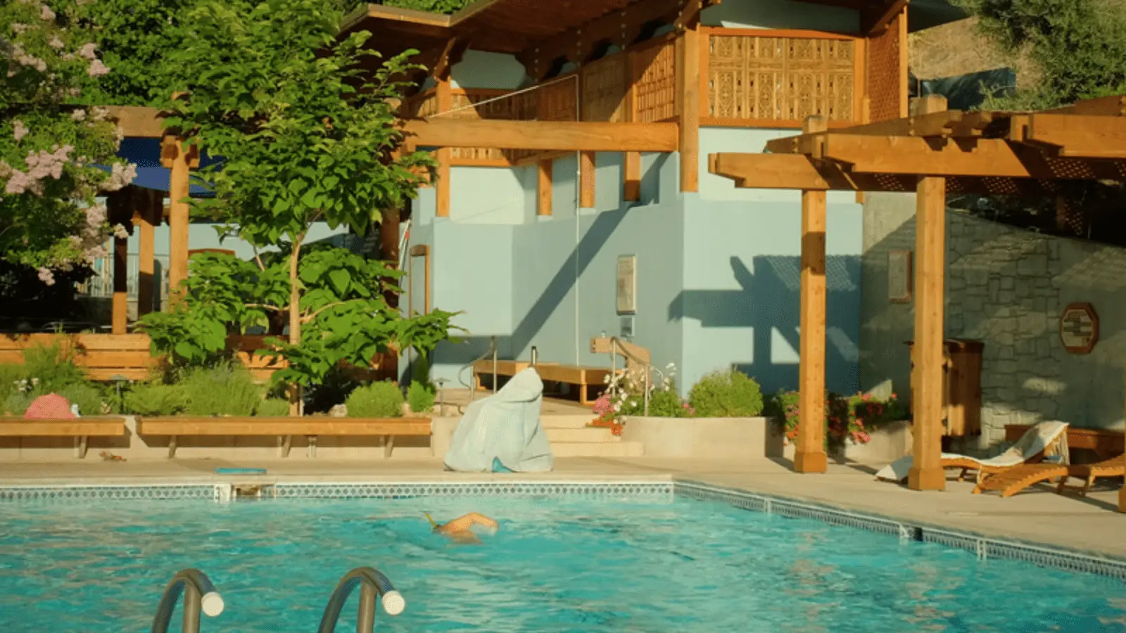 A serene outdoor swimming pool with a person swimming in it. The pool area, reminiscent of hot springs in Northern California, is surrounded by a wooden deck with potted plants and comfortable seating. In the background, there is a charming building with blue walls and a wooden pergola providing shade.