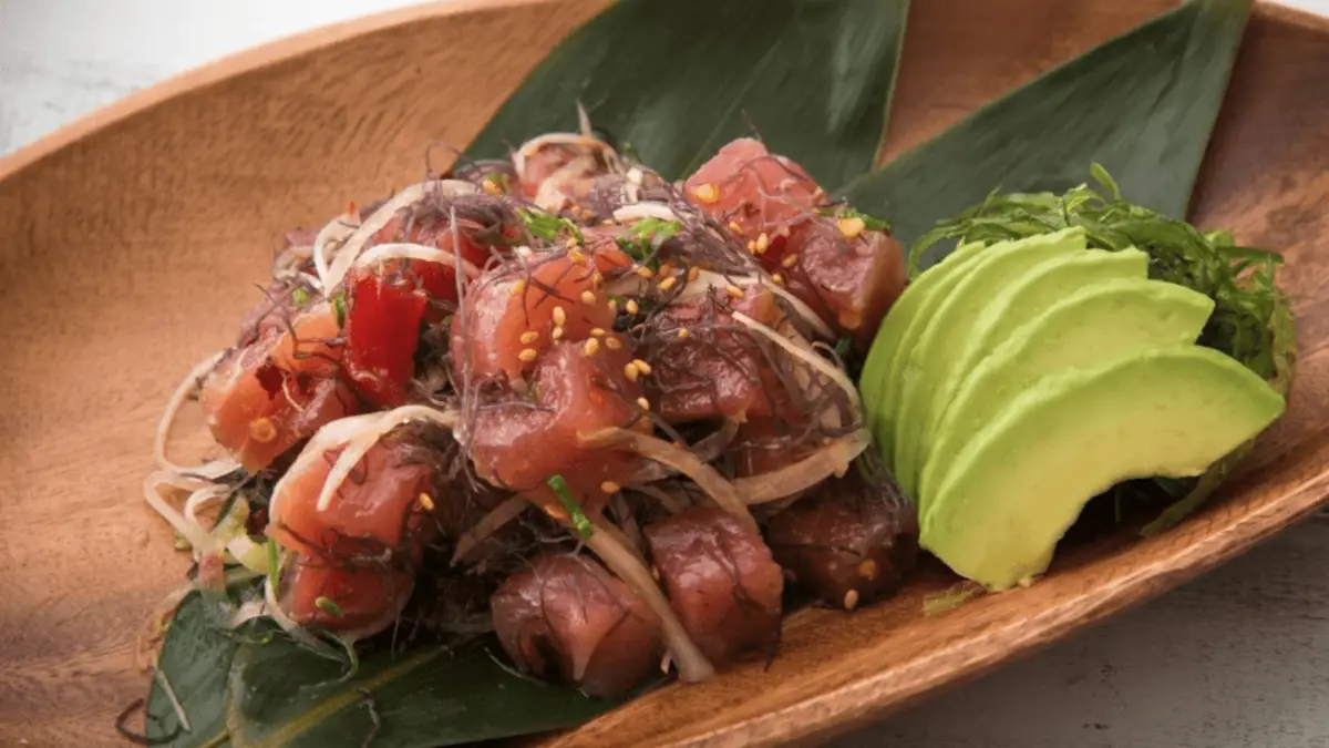 Wooden plate containing a dish made of cubed raw fish mixed with thinly sliced onions and seaweed, garnished with sesame seeds and spices. On the side, there are slices of ripe avocado. This quintessential Hawaiian produce is presented on a green leaf.