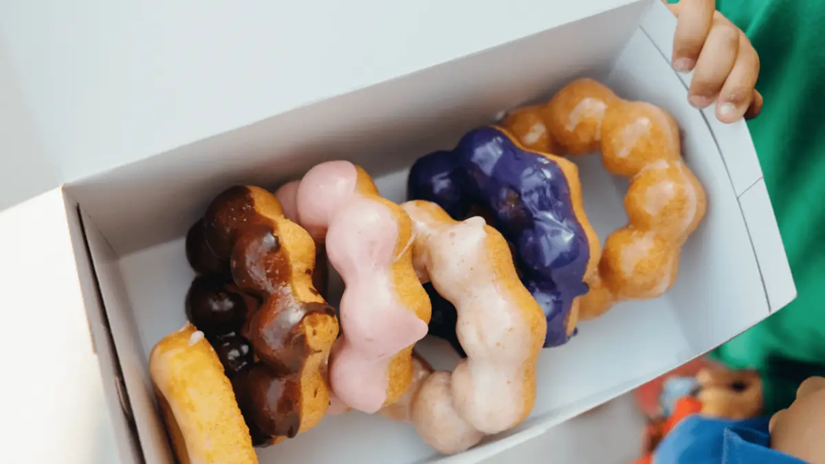 A child's hand is visible holding a box of five donuts with distinctive bumpy textures and ring shapes, each glazed in vibrant colors: chocolate, pink, purple, light pink, and plain. For those seeking the best desserts Maui Wailea has to offer, these delightful treats are a must-try.