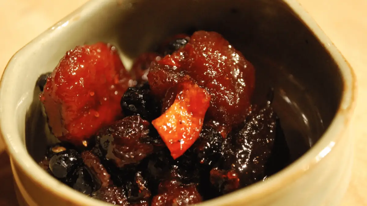 A small ceramic bowl filled with a sticky, glistening mixture of red and dark brown chunks, including pieces of fruit and what appear to be beans or seeds. This dish, reminiscent of the condiments that define Hawaiian culture, looks like it might be a type of preserved or fermented food.