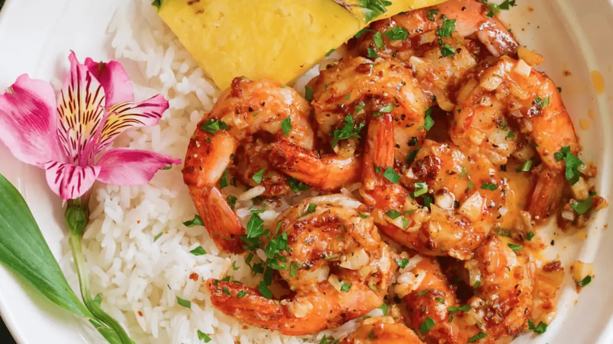 A bowl of white rice topped with succulent, garlic butter shrimp garnished with chopped parsley. A slice of pineapple and a pink flower are placed beside the seafood, adding vibrant color to the dish, echoing the foods that define Hawaiian culture.