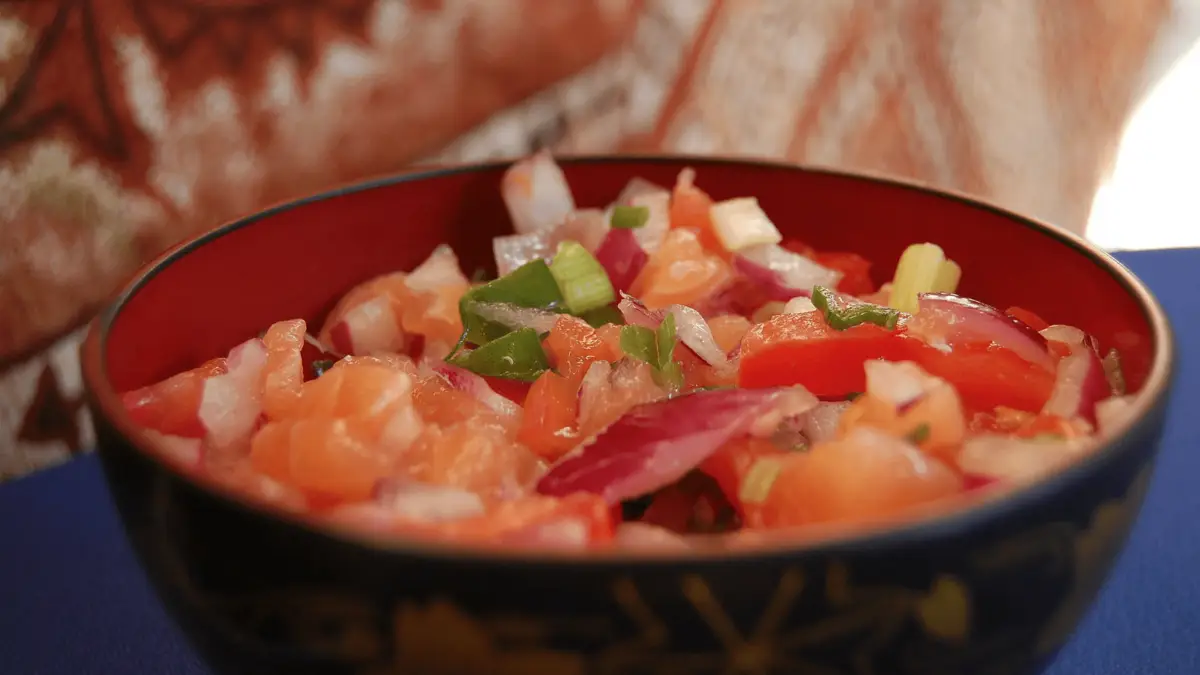 A close-up of a colorful bowl of ceviche containing diced raw fish, red onions, green onions, and tomatoes. The background shows a blurry, warmly lit cloth or tapestry, and the bowl is placed on a blue surface. This dish is among the seafood delights that define Hawaiian cuisine.