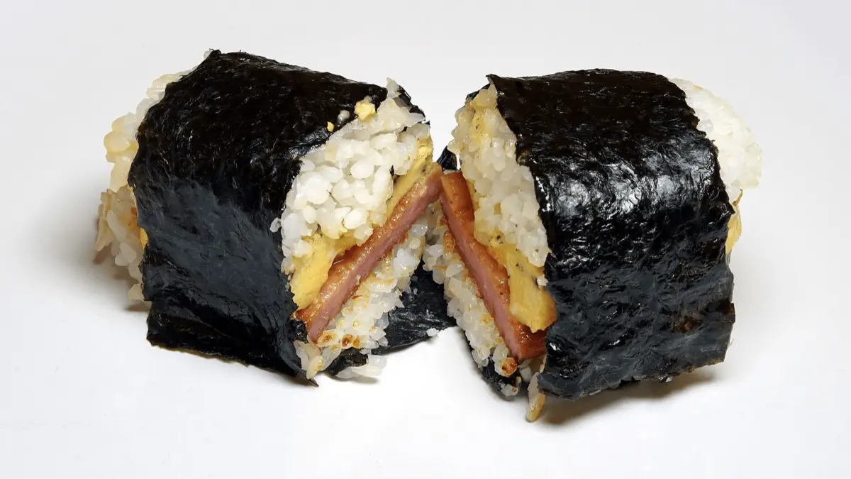 A sushi roll cut in half, revealing layers of white rice, cooked eggs, and a slice of Spam in the middle—one of the foods that define Hawaiian culture—all wrapped in a sheet of black seaweed. The sushi is placed on a plain white background.