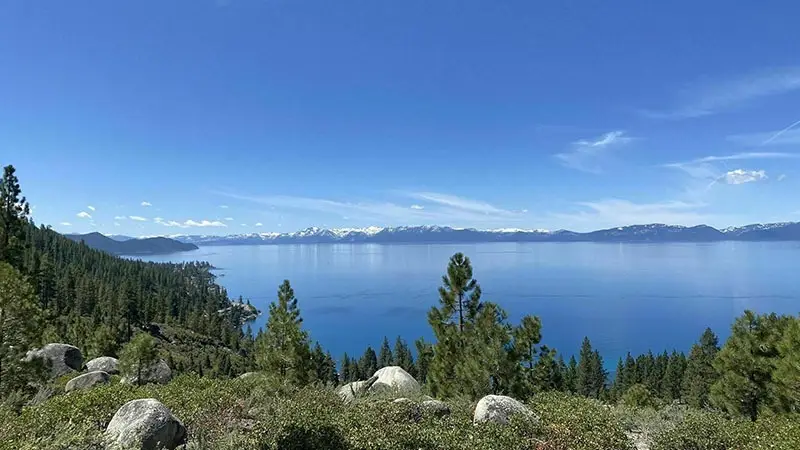 A scenic view of a large, calm lake surrounded by lush green forest and rocky terrain under a clear blue sky. Snow-capped mountains are visible in the distance, enhancing the serene and picturesque landscape—perfect for those seeking local getaways.