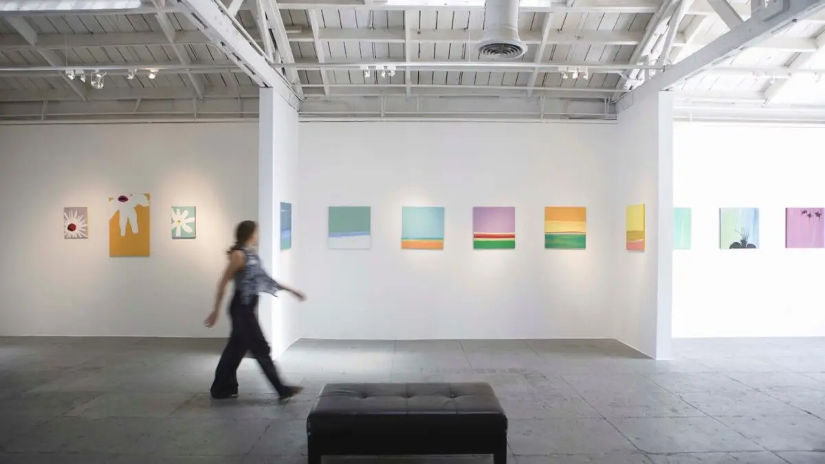 A person on a Napa Valley visit strolls past colorful abstract paintings displayed on the walls of a bright, modern art gallery with a high ceiling. A black bench is positioned in the foreground on the minimalistic concrete floor.